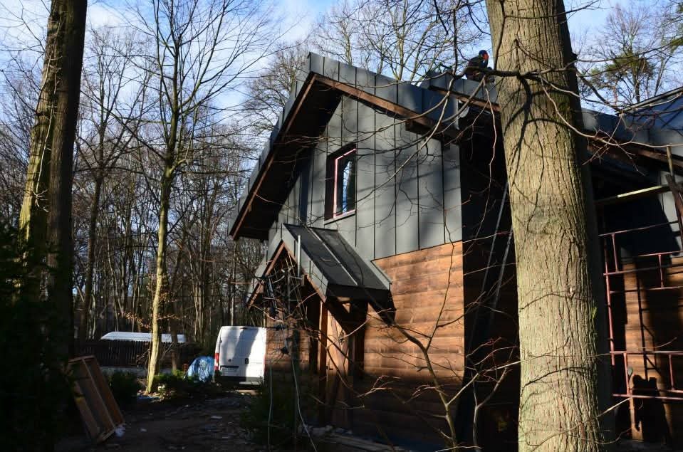 Dom z ciemną, metalową elewacją i dachem, otoczony drzewami. Widoczny fragment rusztowania i pracownik na dachu. Nietypowe połączenie materiałów elewacyjnych.