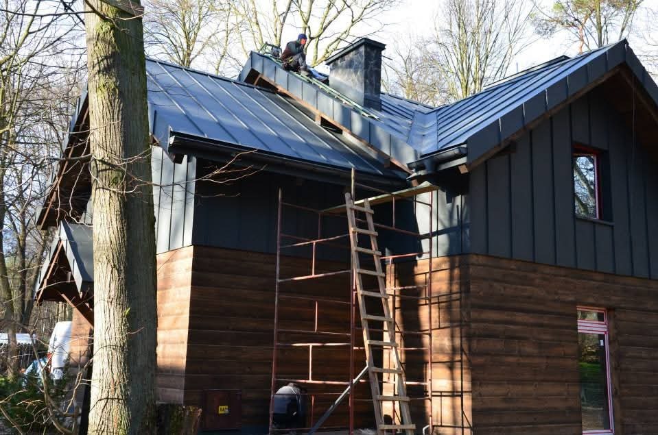 Dom z ciemną, metalową blachą na dachu i drewnianą elewacją. Na dachu pracuje dekarz, widoczne rusztowania i komin. Ujęcie z dołu, drzewo na pierwszym planie.