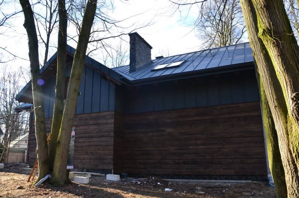 Nowoczesny dom z ciemną blachą na dachu i drewnianą elewacją. Widoczne okna dachowe i komin z kamienia. Ujęcie z dołu, drzewa na pierwszym planie.