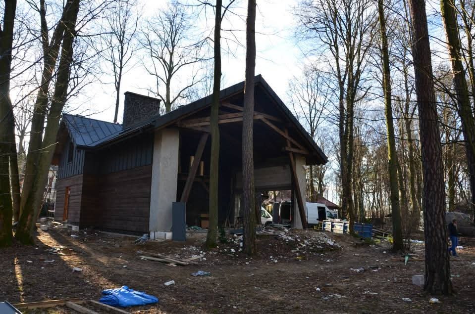 Budynek w lesie z widoczną konstrukcją dachu i kominem z kamienia. Częściowo wykończona elewacja z drewnianych paneli. Prace w toku, widoczne materiały budowlane.