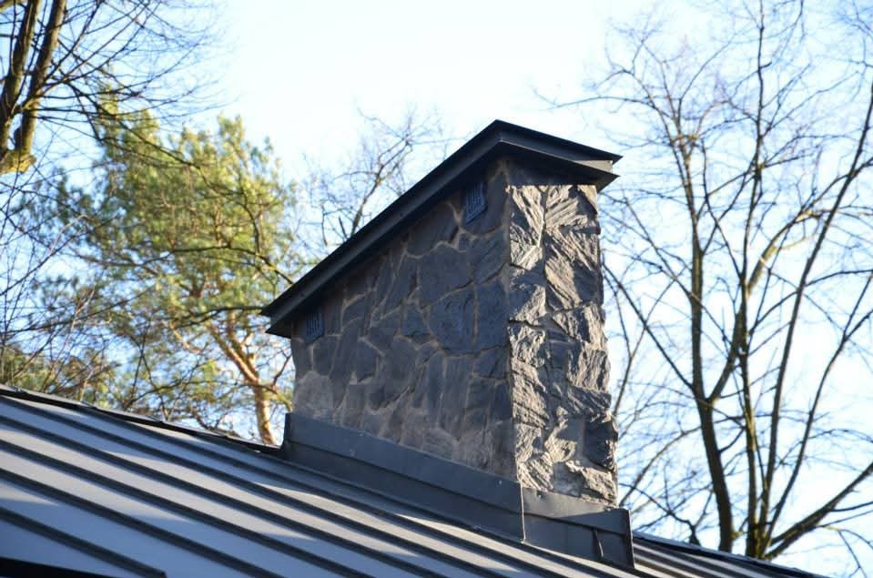 Szary dach z blachy trapezowej z kamiennym kominem w słoneczny dzień. Widoczne drzewa bez liści w tle.