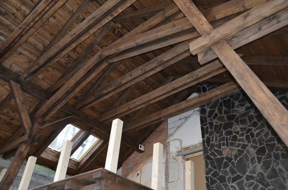 Drewniana więźba dachowa z widocznymi elementami konstrukcyjnymi i oknami dachowymi, fragment ściany z kamienia w tle. Widoczne elementy rusztowania.