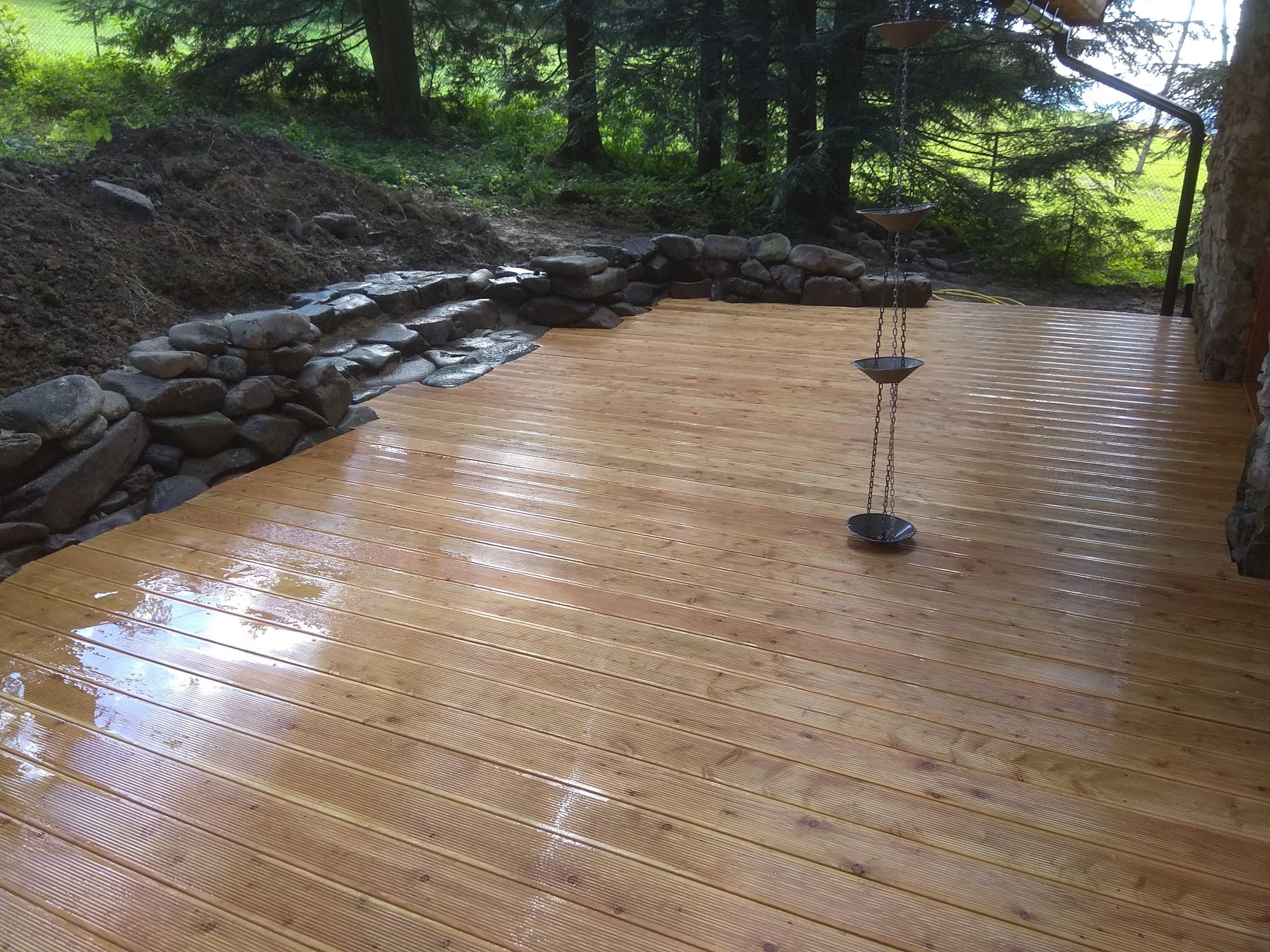 Nowy, drewniany taras z desek ryflowanych, z kamiennym obrzeżem i łańcuchem deszczowym. Widok na ogród z drzewami iglastymi w tle. Świeżo po deszczu, mokra powierzchnia.