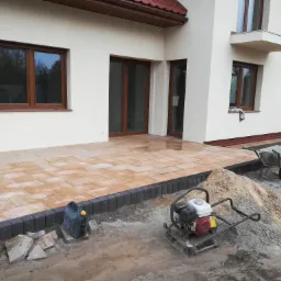 Układanie kostki brukowej na tarasie przylegającym do domu, widoczny ubijak wibracyjny Honda i taczka z piaskiem.