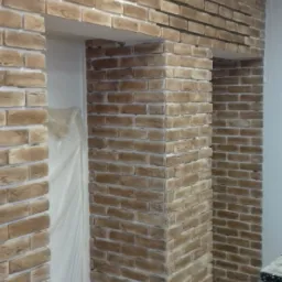 Ściana wykończona cegłą dekoracyjną w odcieniach beżu i brązu, widoczne fragmenty drzwi zabezpieczonych folią oraz drabina malarska.