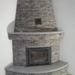 Obudowa kominka z szarego kamienia łupanego, z półkami z szarego granitu, wykończona w narożniku pomieszczenia, z czarnym wkładem kominkowym.