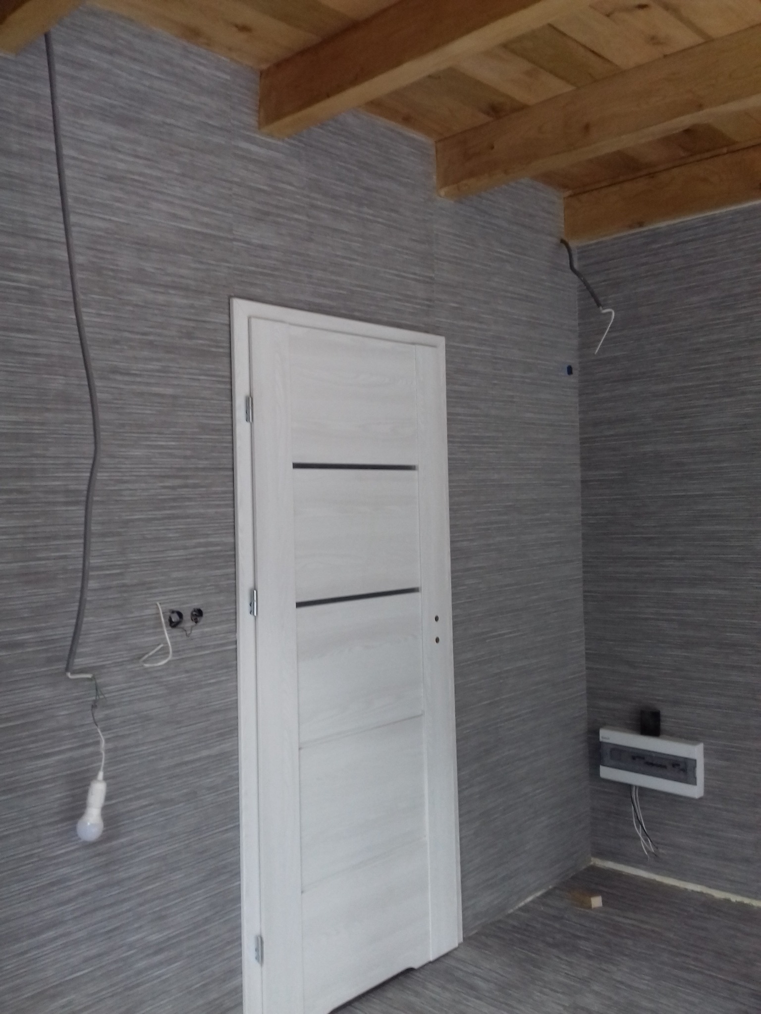 Wnętrze w trakcie wykończenia: szara ściana z poziomym wzorem, białe drzwi z trzema poziomymi wgłębieniami, drewniany sufit, wisząca żarówka i przewody elektryczne.
