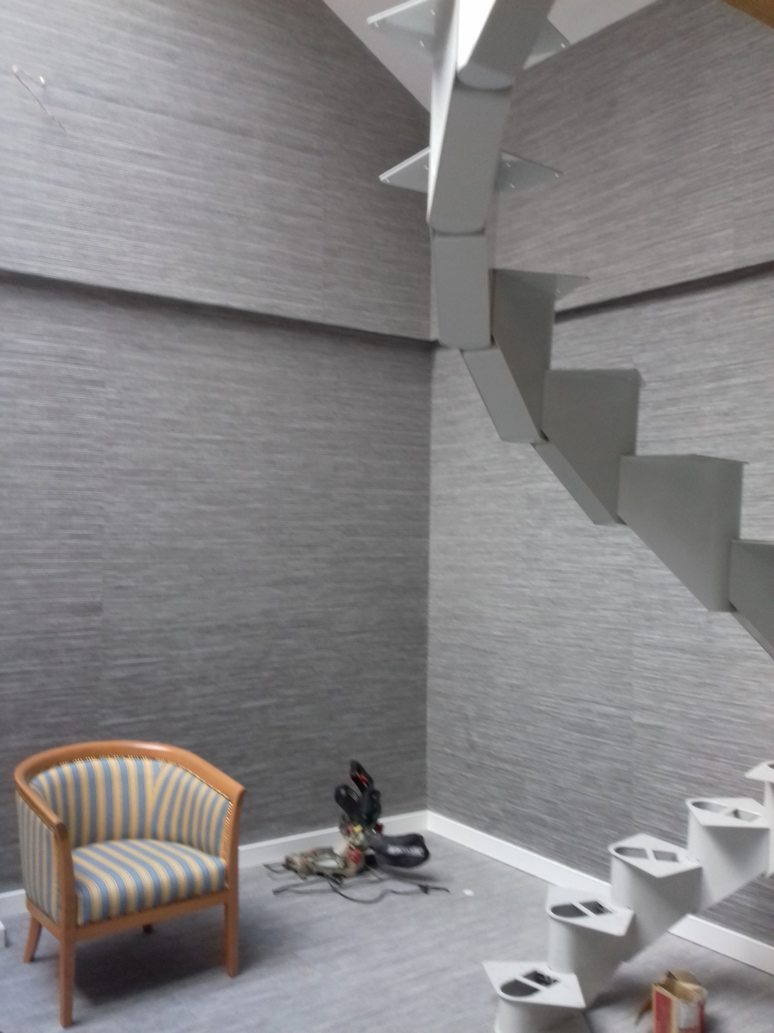 Pokój z szarymi ścianami o teksturze tkaniny, nowoczesne schody metalowe bez poręczy, fotel w niebiesko-żółte paski i narzędzia budowlane na podłodze.