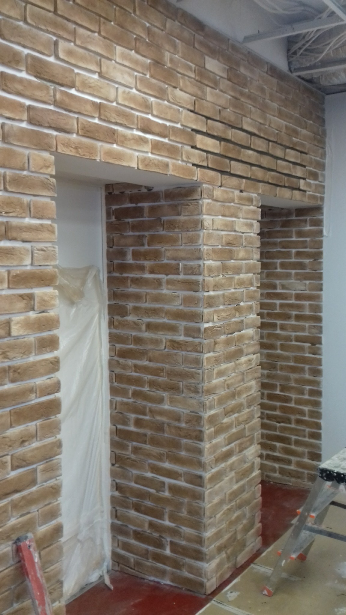 Ściana wykończona cegłą dekoracyjną w odcieniach beżu i brązu, widoczne fragmenty drzwi zabezpieczonych folią oraz drabina malarska.