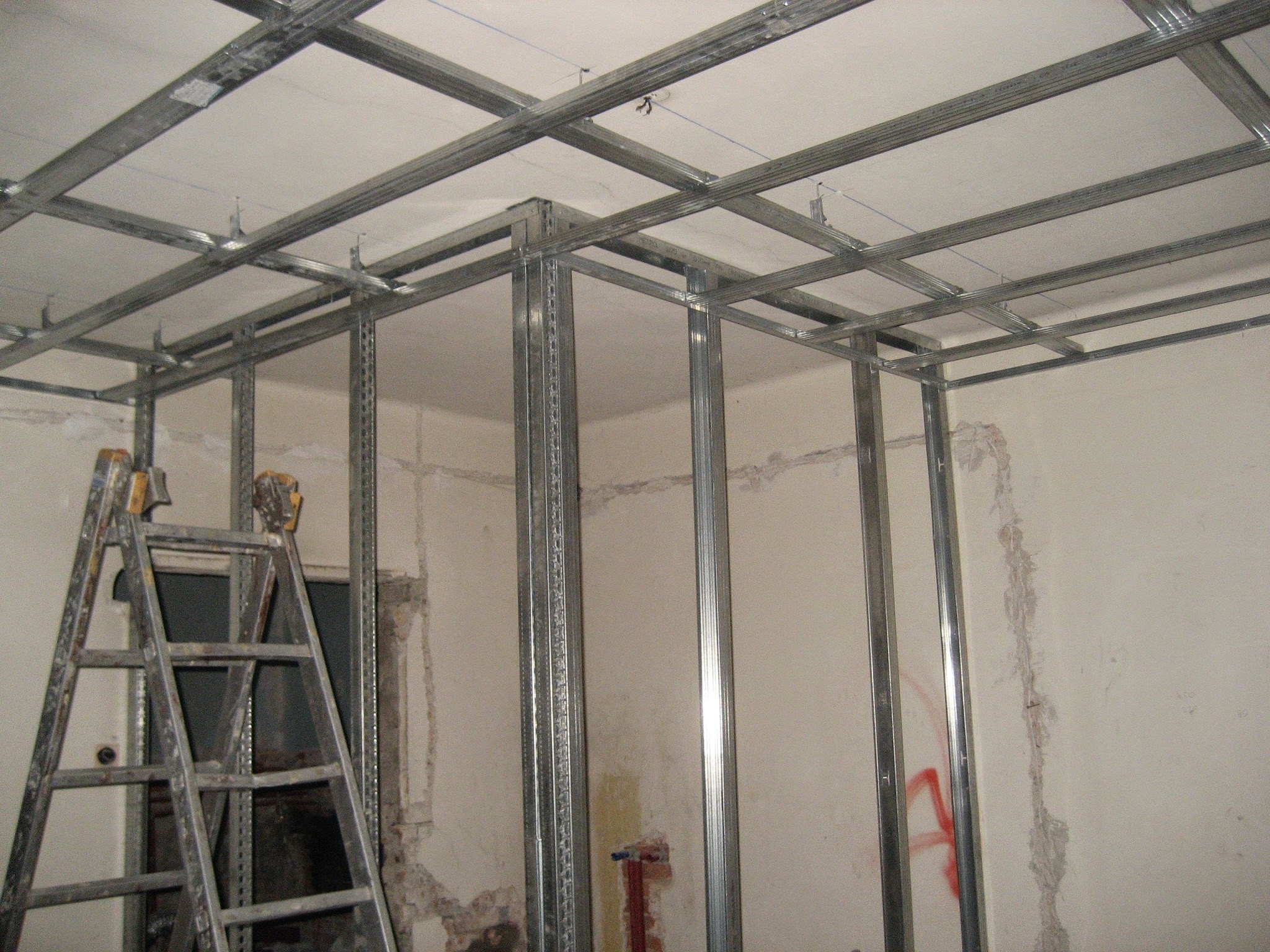 Metalowa konstrukcja sufitu podwieszanego i ścianki działowej, widoczna drabina i fragmenty starego muru, etap prac wykończeniowych.