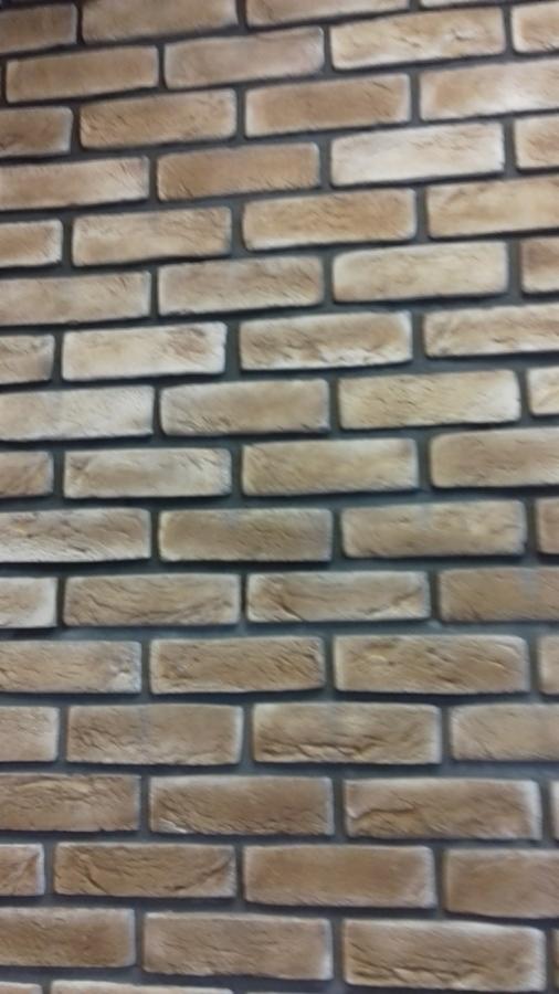 Ściana wyłożona dekoracyjną cegłą w odcieniach beżu i brązu z ciemnoszarą fugą, ułożona pod kątem, z widoczną fakturą cegieł.
