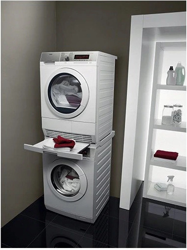 Kompaktowa pralko-suszarka ustawiona jedna na drugiej w nowoczesnej łazience z czarną podłogą i białymi półkami z detergentami.