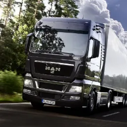 Czarny ciągnik siodłowy MAN z białą naczepą w dynamicznym ujęciu na tle leśnej drogi i błękitnego nieba z cumulusami.