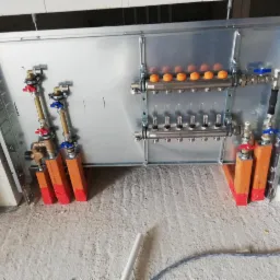 Rozdzielacz ogrzewania podłogowego zamontowany w metalowej szafce, z widocznymi zaworami, rurami i elementami mocującymi, w surowym stanie wykończeniowym pomieszczenia.