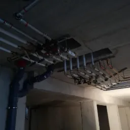 Skomplikowany układ rur instalacyjnych pod betonowym sufitem, widoczne rury z tworzywa, miedziane i metalowe, liczne zawory i złączki, fragment konstrukcji budynku w tle.