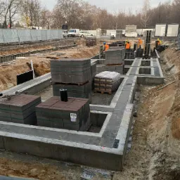 Budowa fundamentów: zalane betonem ławy fundamentowe z ułożonymi bloczkami betonowymi na paletach, widoczni pracownicy w pomarańczowych kamizelkach.