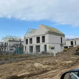 Wznoszone domy z białego bloczka, widoczne więźby dachowe, teren budowy z ogrodzeniem i stertą palet drewnianych.