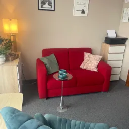 Kącik terapeutyczny z czerwonym dwuosobowym siedziskiem, stolikiem z dwiema filiżankami, niebieskim fotelem na pierwszym planie oraz dekoracjami ściennymi w gabinecie.