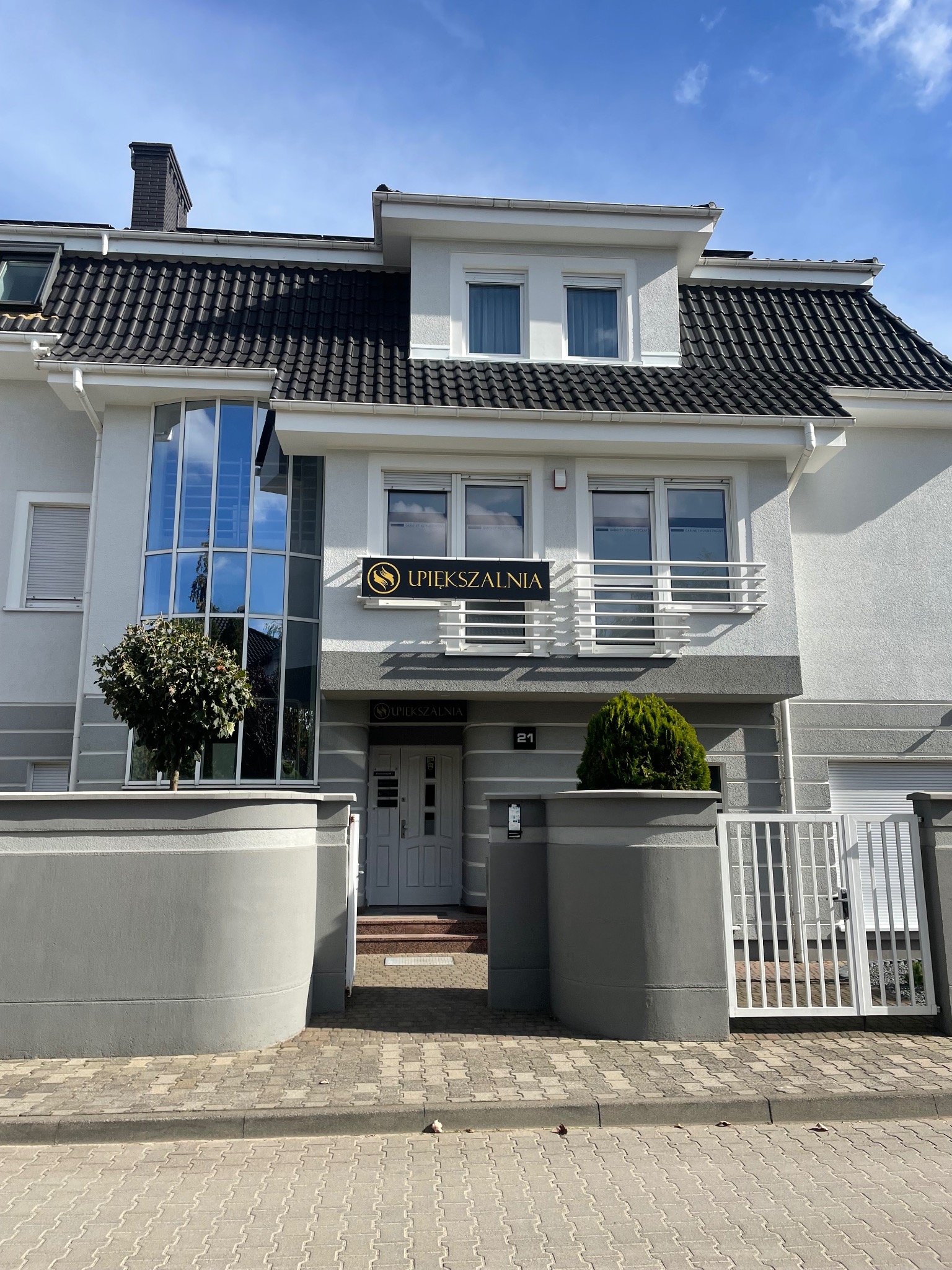 Elewacja budynku z szyldem gabinetu kosmetycznego, widoczne okna, krzewy i numer 21.