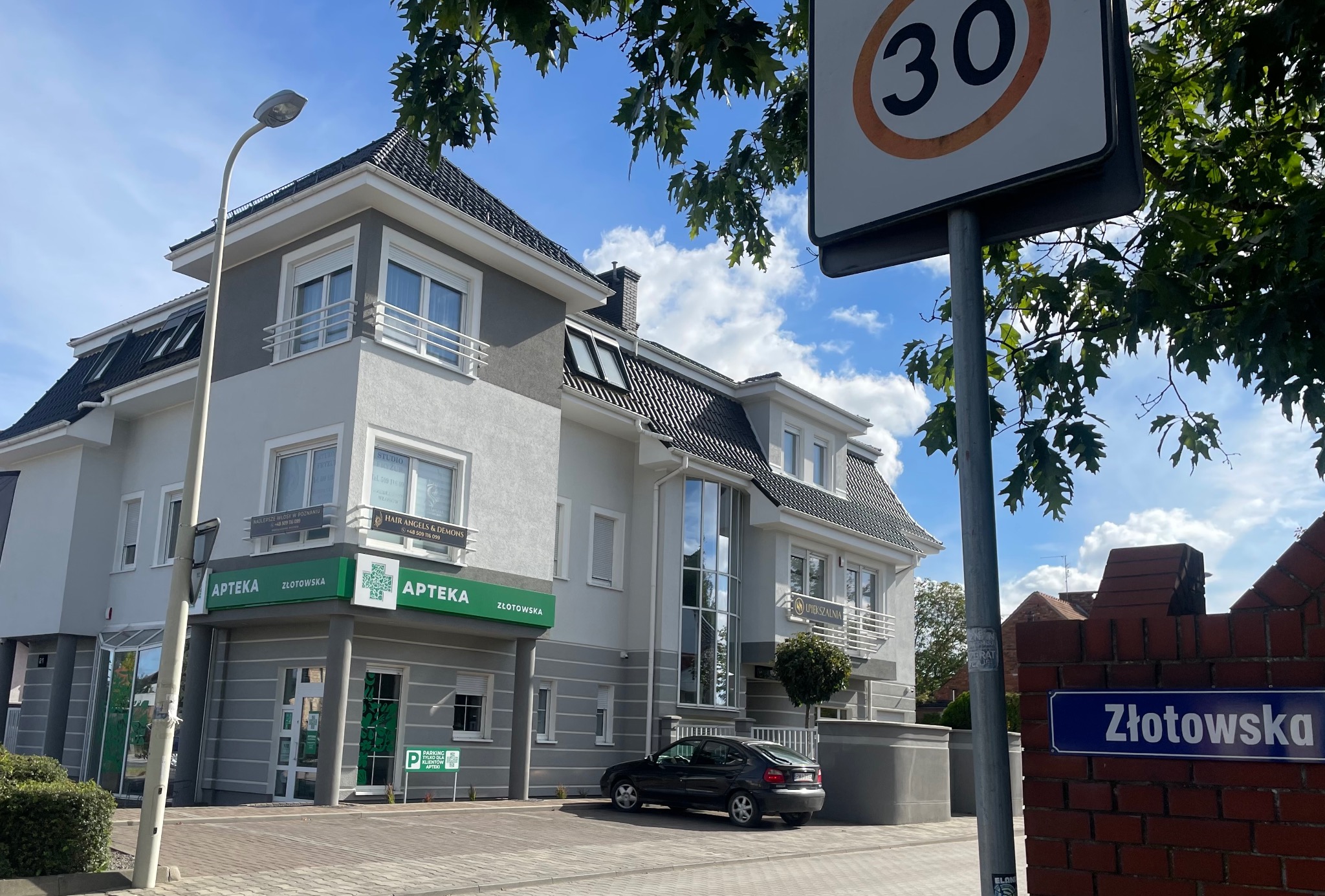 Budynek apteki Złotowska z zielonym logo i napisem, widok z ulicy, czarny samochód zaparkowany przed budynkiem, znak drogowy z ograniczeniem prędkości do 30 km/h.