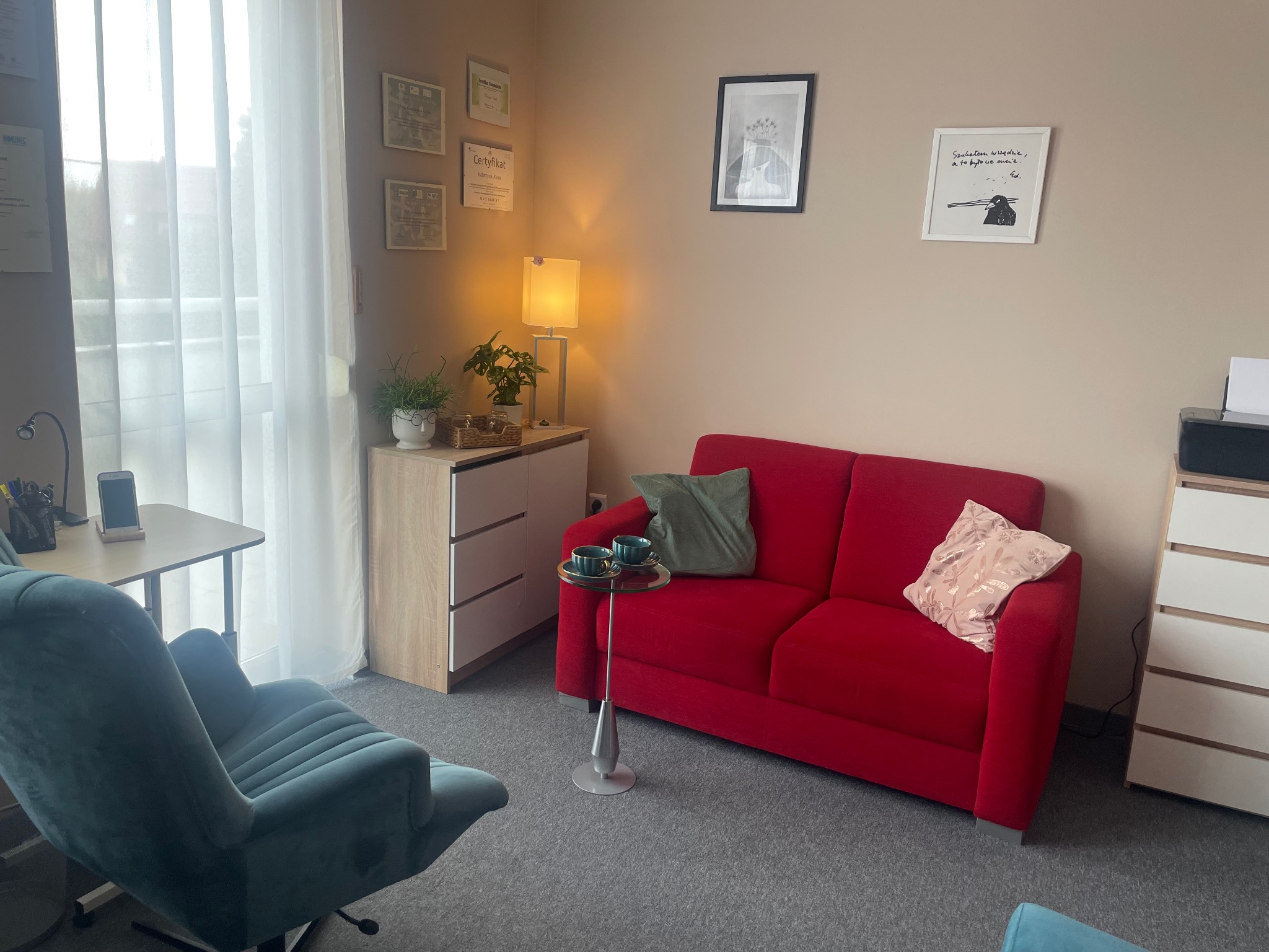 Jasne, przytulne wnętrze gabinetu z czerwonym dwuosobowym siedziskiem, zielonymi poduszkami, stolikiem z filiżankami, zielonym fotelem na kółkach, biurkiem i komodą, ozdobione certyfikatami...