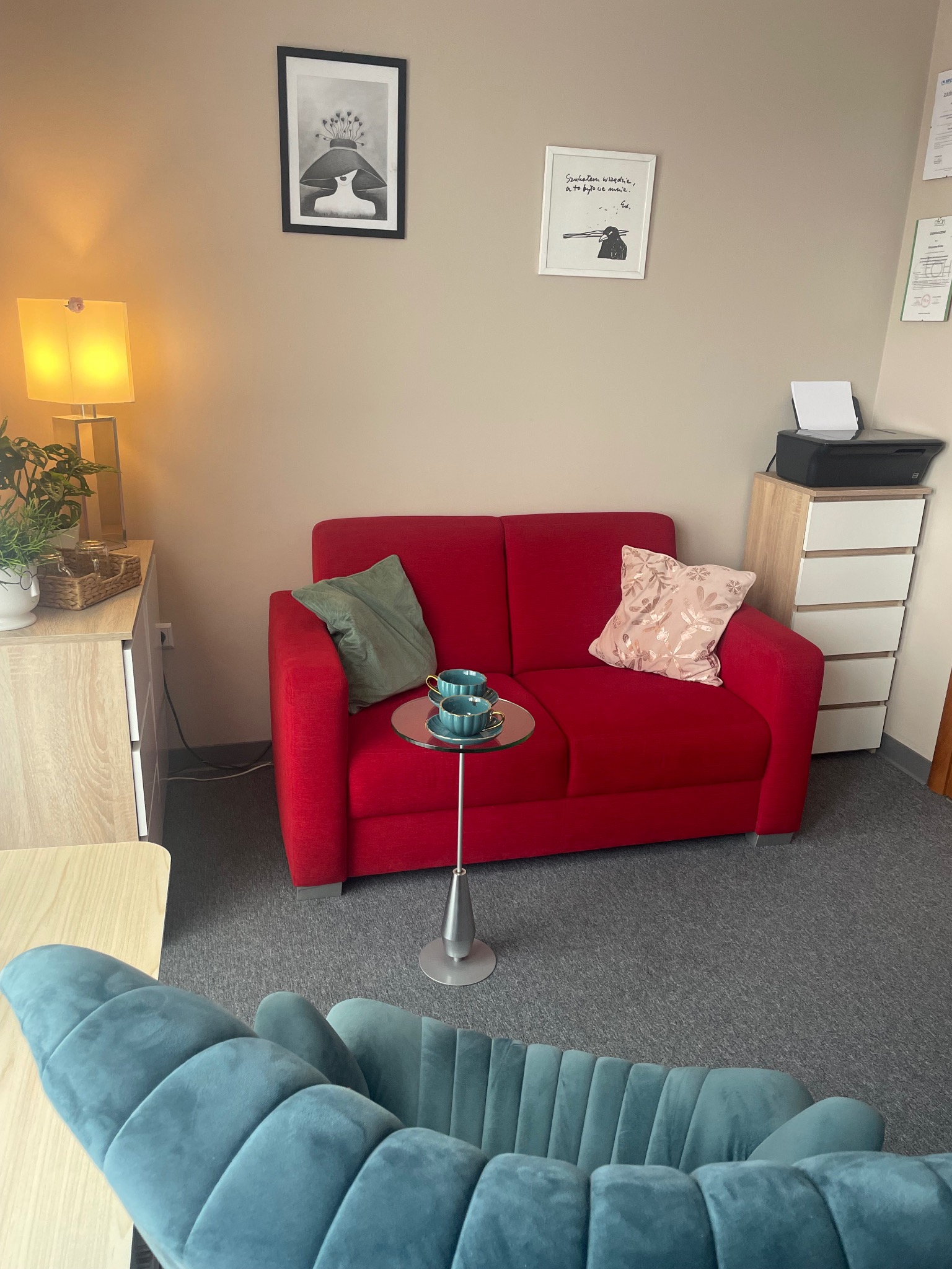Kącik terapeutyczny z czerwonym dwuosobowym siedziskiem, stolikiem z dwiema filiżankami, niebieskim fotelem na pierwszym planie oraz dekoracjami ściennymi w gabinecie.