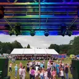 Plenerowa impreza masowa z dużą sceną oświetloną reflektorami, białym namiotem cateringowym w tle i tłumem ludzi na trawniku.