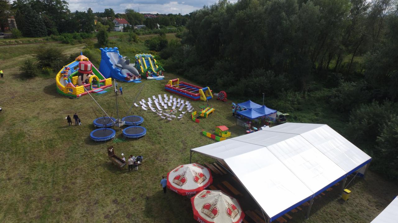 Widok z góry na rozłożoną strefę rekreacyjną na trawiastym terenie: dmuchane zamki, trampoliny, boisko do piłkarzyków, leżaki i biały namiot z parasolami Tyskie.