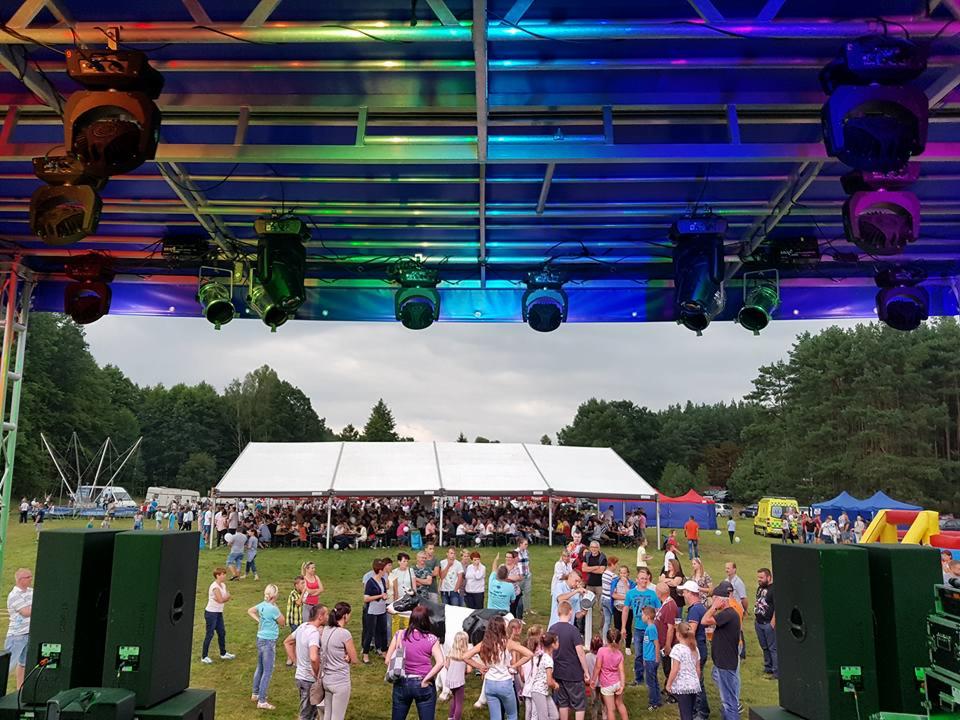 Plenerowa impreza masowa z dużą sceną oświetloną reflektorami, białym namiotem cateringowym w tle i tłumem ludzi na trawniku.