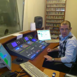 Operator siedzi przed konsoletą mikserską z ekranami i suwakami w studiu nagrań, w tle regał z płytami CD.