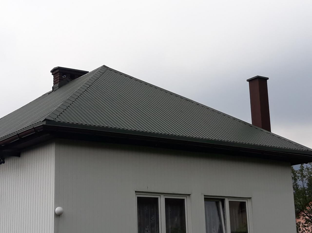 Dom z nowym, ciemnozielonym dachem z blachy trapezowej, brązowymi rynnami i dwoma kominami na tle pochmurnego nieba.