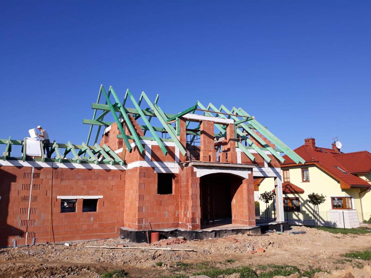 Budowa domu jednorodzinnego w trakcie wznoszenia więźby dachowej z zielonych belek, widoczny mur z czerwonej cegły i pracownik na dachu, błękitne niebo w tle.