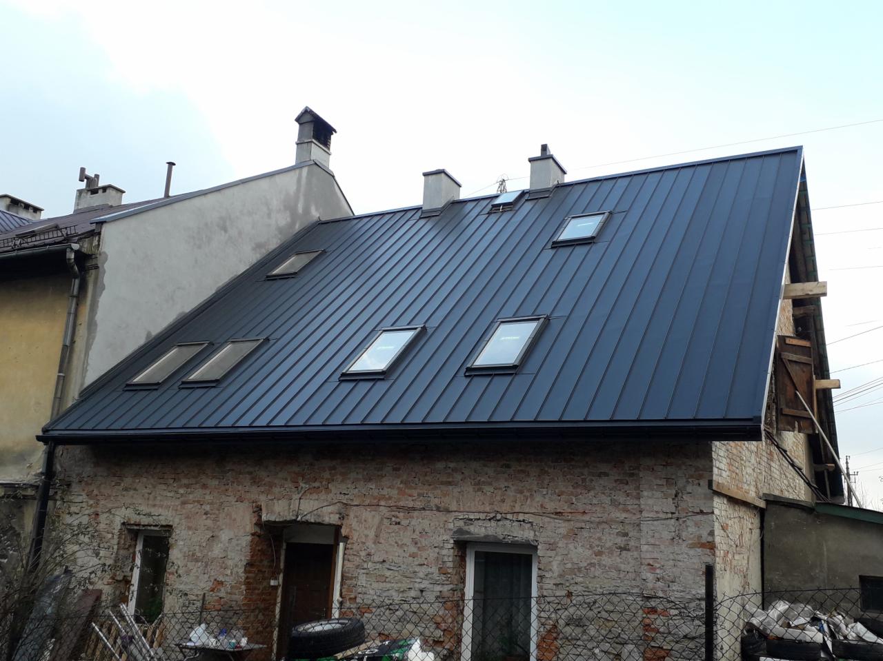 Nowy, ciemny dach z blachy trapezowej z oknami dachowymi na starym, ceglanym budynku z widocznymi śladami renowacji.