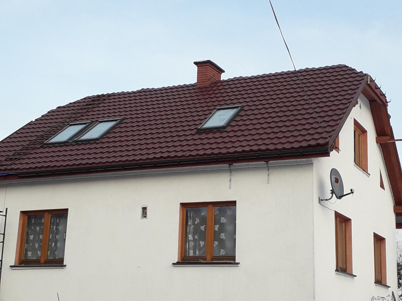 Dom jednorodzinny z nowym, bordowym dachem z blachodachówki, trzema oknami dachowymi i ceglanym kominem. Biała elewacja z drewnianymi oknami i anteną satelitarną.
