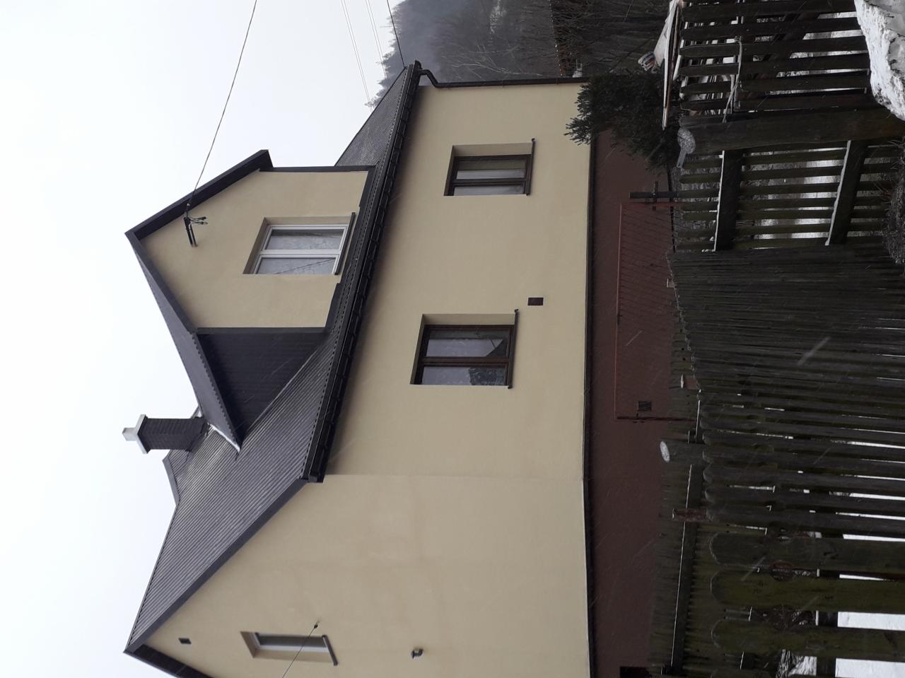 Dwupiętrowy dom z ciemnym dachem i beżową elewacją, widoczny z niskiego kąta, przysłonięty częściowo przez drewniany płot. Na dachu widoczny komin.