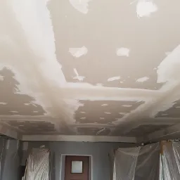 Sufit z płyt kartonowo-gipsowych w trakcie szpachlowania, widoczne łączenia i miejsca po wkrętach wypełnione gładzią, profile metalowe pod sufitem, zabezpieczone meble folią malarską.