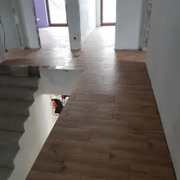 Układanie brązowych płytek imitujących drewno na podłodze w nowym domu, widoczne schody w surowym stanie i niedokończone ściany.