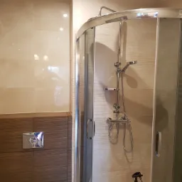 Nowoczesna kabina prysznicowa z chromowanymi elementami, beżowe płytki na ścianach, widoczny panel z brązowymi płytkami i chromowanym przyciskiem spłuczki, oświetlenie punktowe.