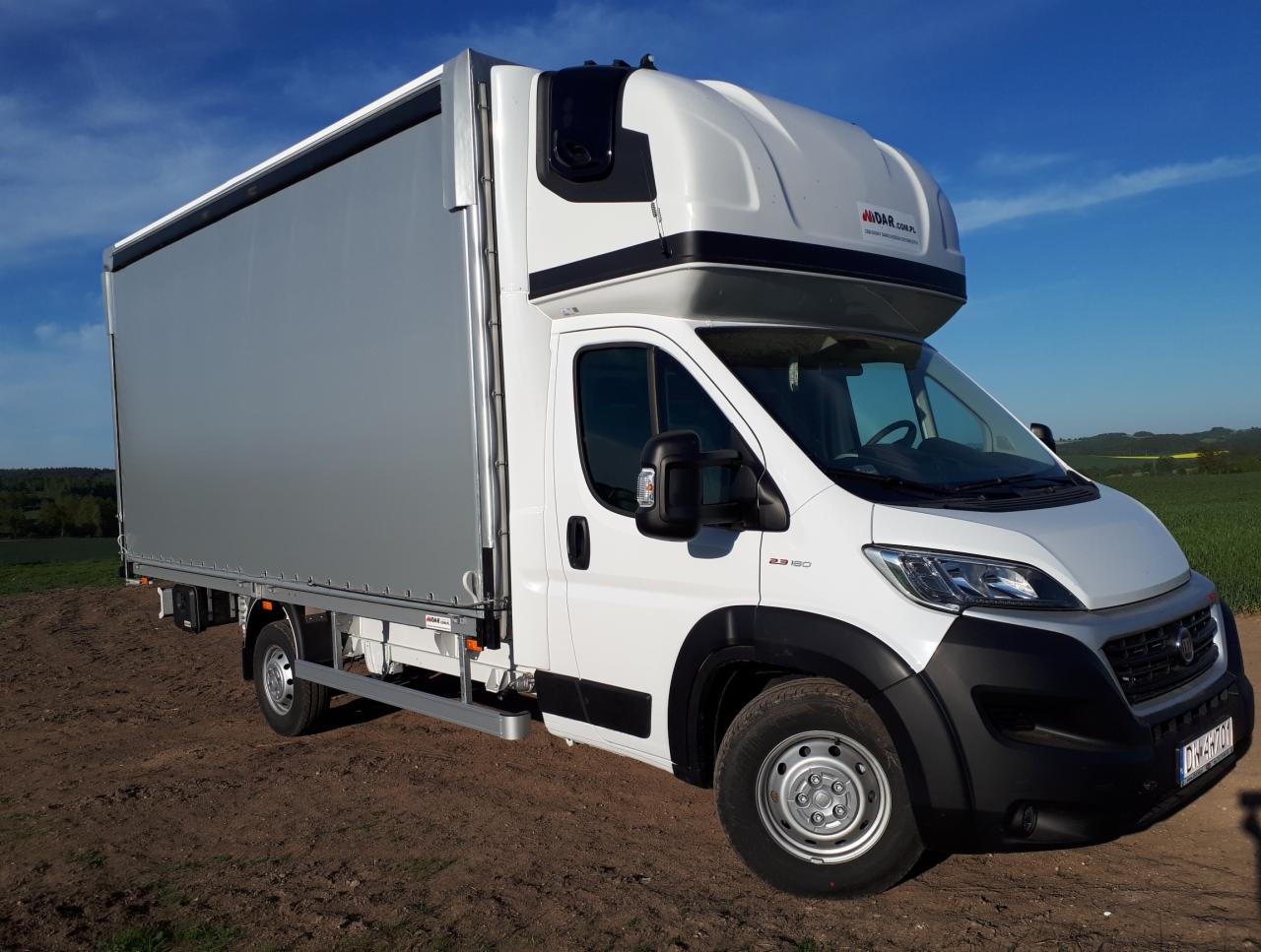 Biały samochód dostawczy z plandeką na tle błękitnego nieba i zielonego pola, widoczny z boku, z logo producenta zabudowy na dachu.