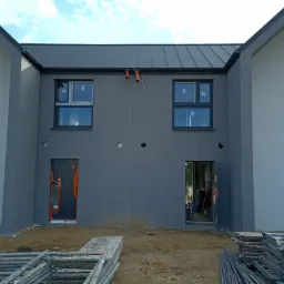 Nowo wykończona elewacja budynku bliźniaczego w szarościach, z widocznymi oknami i przygotowanymi otworami drzwiowymi, z których jeden jest zabezpieczony folią, a drugi otwarty, z widocznym...