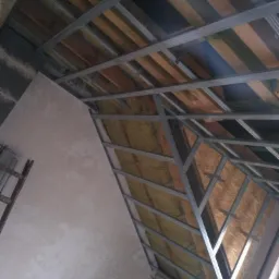 Montaż metalowej konstrukcji sufitu podwieszanego na poddaszu, widoczna wełna mineralna jako izolacja, ściana w stanie surowym.