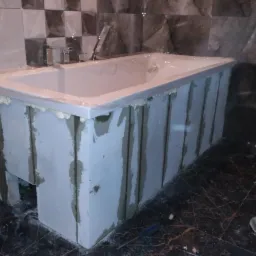 Wanna obudowywana płytami, widoczna pianka montażowa, ciemne marmurowe płytki na ścianie i podłodze, etap prac wykończeniowych w łazience.