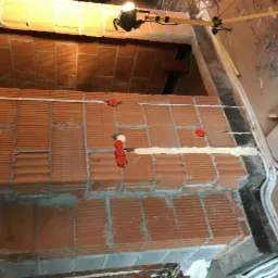 Wewnętrzna ściana z cegły z widocznymi przewodami elektrycznymi w białej listwie, czerwone puszki instalacyjne i żółty statyw oświetleniowy w tle.