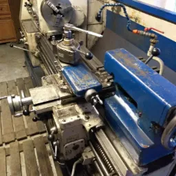 Używana tokarka z uchwytem tokarskim i narzędziem skrawającym, widoczna instalacja chłodzenia i narzędzia na obudowie.