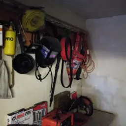 Warsztatowy kąt z narzędziami: maska spawalnicza, butla gazowa, przewody, ładowarka akumulatorów i pilarka łańcuchowa.