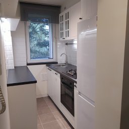 REN-BUD.WAW KRZYSZTOF GRAB - Wnętrze małej kuchni w bieli z czarnymi akcentami, widok na okno z roletą w szarym odcieniu, białe szafki z czarnymi uchwytami, czarny blat i płyta grzewcza, brązowe płytki podłogowe.