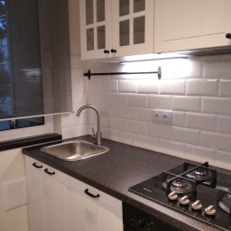 REN-BUD.WAW KRZYSZTOF GRAB - Fragment kuchni z białymi szafkami z przeszklonymi frontami, czarnym blatem, metalowym zlewem i płytą gazową, oświetlony paskiem LED pod szafkami.