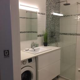 REN-BUD.WAW KRZYSZTOF GRAB - Nowoczesna łazienka z białą zabudową umywalki, pod którą umieszczono pralkę; kabina prysznicowa ze szklaną ścianą i mozaiką na ścianie; oświetlenie LED nad lustrem.