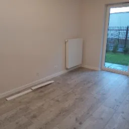 Pokój po remoncie z panelami podłogowymi imitującymi drewno, białymi ścianami, grzejnikiem i drzwiami balkonowymi z widokiem na ogród.