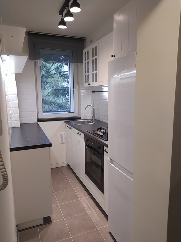 Wnętrze małej kuchni w bieli z czarnymi akcentami, widok na okno z roletą w szarym odcieniu, białe szafki z czarnymi uchwytami, czarny blat i płyta grzewcza, brązowe płytki podłogowe.